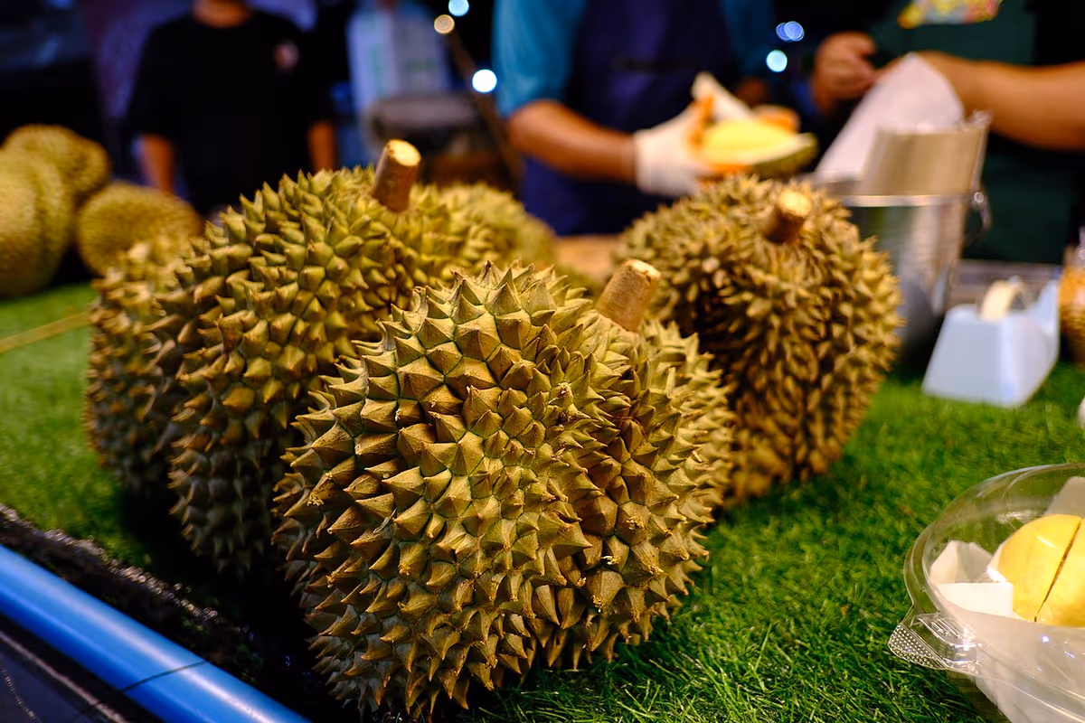 Plusieurs durians épineux présentés sur un étal vert de marché, un vendeur découpant des fruits à l'arrière