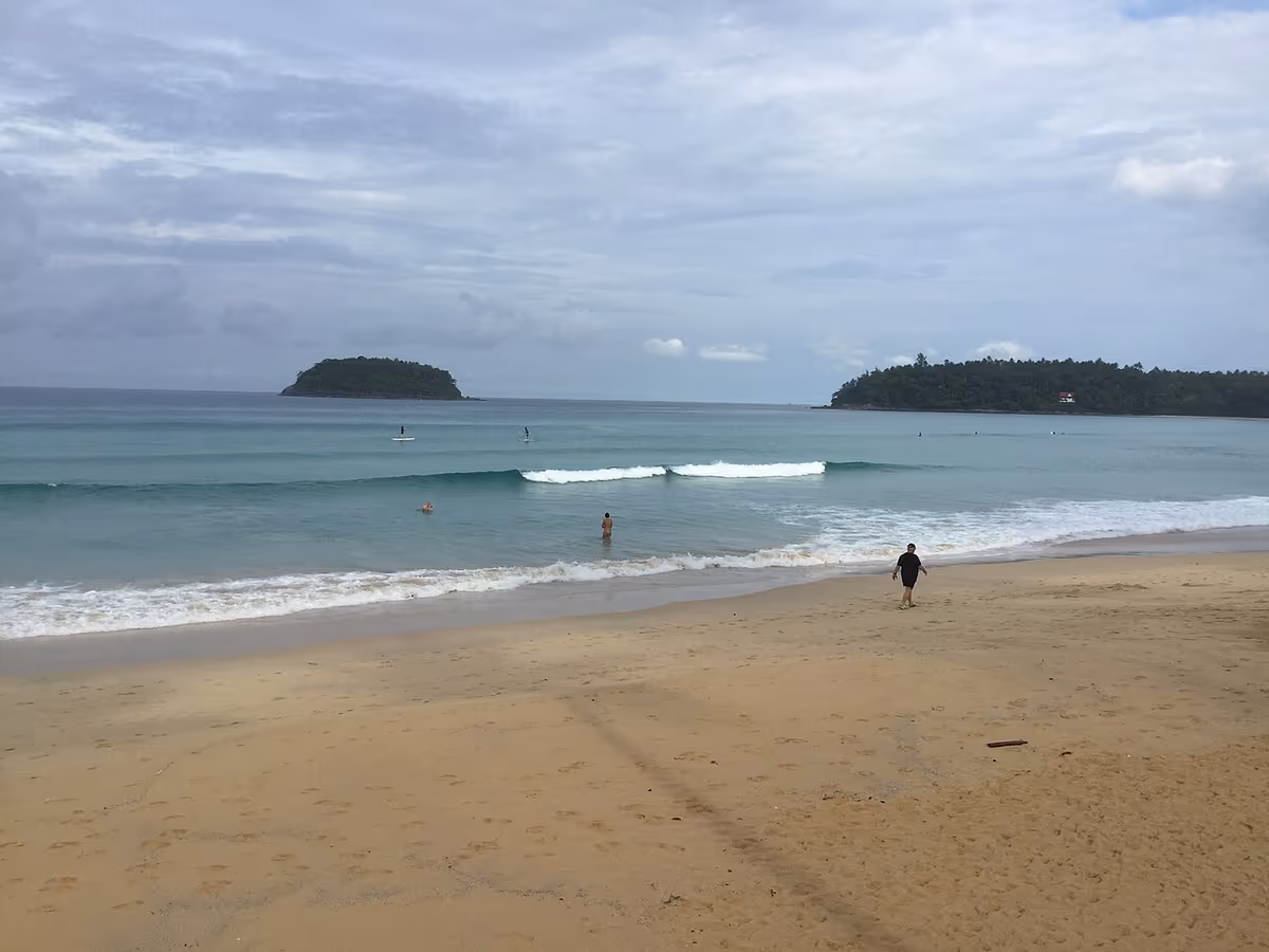 Spiaggia di Kata a Phuket: riva sabbiosa, acqua turchese, onde oceaniche, isole boscose sullo sfondo, luce mattutina.